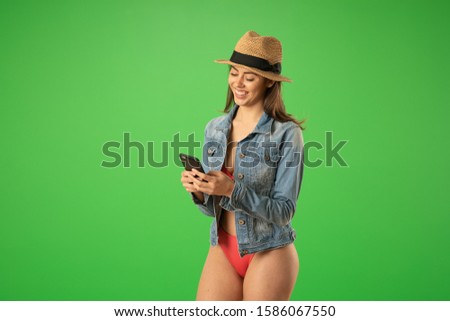 Portrait of cheerful young woman in bikini and jean jacket laughing at text message. Beautiful girl in fedora messaging her friends on sunny beach on greenscreen
