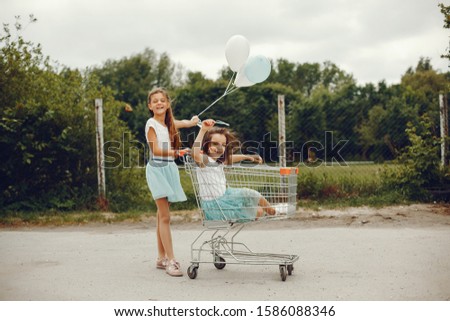 two cute little girls in white T-shirts and blue skirts play in the summer park with balloons and ride in the grocery cart