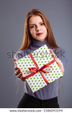 Young caucasian lady with long hair is giving a gift box to you and me isolated on a gray background..