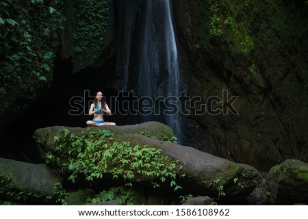 Yoga pose. Young Caucasian woman sitting on the rock, meditating, practicing yoga, pranayama at waterfall. Hands in namaste mudra. Yoga retreat. Leke Leke waterfall, Bali, Indonesia