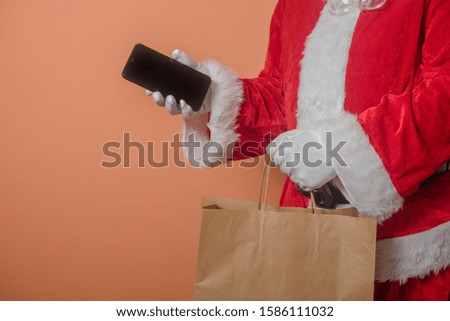 Closeup on a busy Father Santa Claus using showing mobile smart phone and holding shopping paper bag. Mockup copy space modern design display festive decoration background photography. Happy New Year!