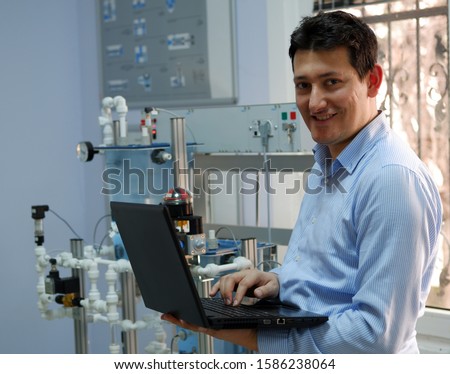  Engineer is programming PLC with his laptop for an analog process kit. Industry 4.0 workshop in an automation education center. Engineer is learning how to program a PLC. Selective focus. Royalty-Free Stock Photo #1586238064