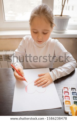 Child making homemade greeting card. A cute little girl paints a heart on a homemade greeting card as a gift. Looking at the camera. Happy valentines day. Art and craft concept. 