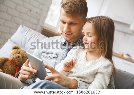Little daughter with dad indoors sitting on the coach browsing digital tablet watching cartoons smiling happy
