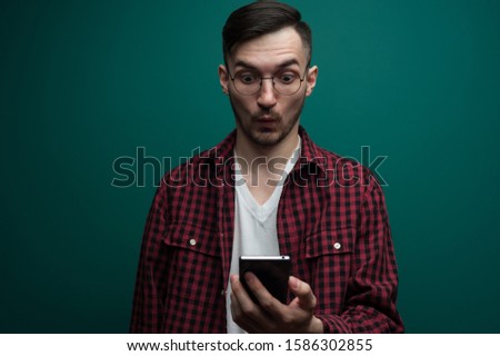 Portrait of an emotional surprised young hipster man in glasses looking at his smartphone on a green background. Concept of discounts in the online store. Advertising space