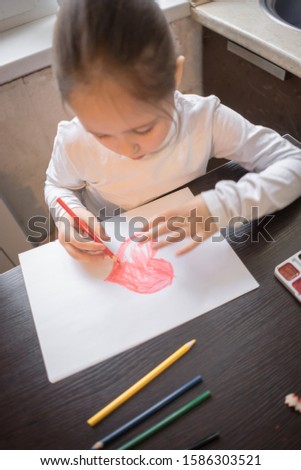Child making homemade greeting card. A cute little girl paints a heart on a homemade greeting card as a gift. Looking at the camera. Happy valentines day. Art and craft concept. 