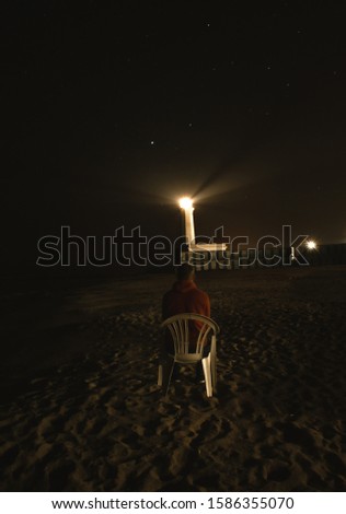 A man sits on a white chair on a sandy beach by the sea and looks towards the burning lighthouse and stars. A young man on a chair sits on the shore and looks at the stars