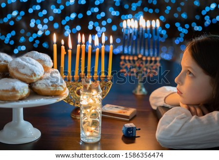 Child girl Looking at Menorah Candles on wooden table and sufganiyot on background light glitter bokeh overlay. Hanukkah jewish holiday Israel hebrew traditional family celebration invitation design
