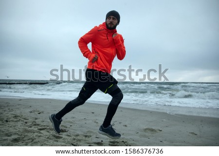 Photo of young bearded man in good physical shape looking over his shoulder while doing morning run at the seaside. Male fitness jogging training wellness concept
