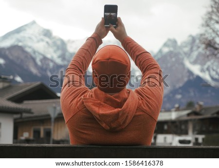 Man taking photos with cell mobile phone on Alps mountains background. Garmisch-Partenkirchen, Germany. winter season.  snowy hills and cloudy sky. male wear orange hoodie and hat. back rear view.