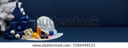 Construction hard hat, color palette guide, paint brushes, Christmas ornaments, fir tree, gifts on dark blue background with copy space. New Year. Christmas. Concept of construction or design office