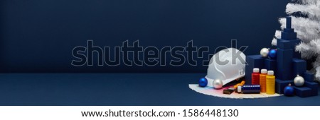 Construction hard hat, color palette guide, paint brushes, Christmas ornaments, white fir tree on dark blue background with copy space. New Year and Christmas. Concept of construction or design office