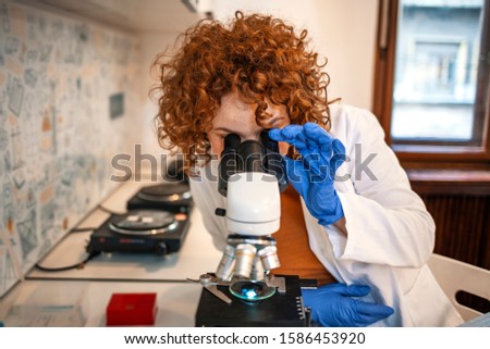 Young scientist looking through a microscope in a laboratory. Young scientist doing some research. Adult caucasian beauty woman chemist looking at microscope against chemistry lab background.