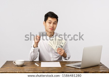 Serious and cheeky asian businessman sitting his office with lots cash, big money, holding credit card and dollars, advice keep money safe, use banking system, consulting client, white background