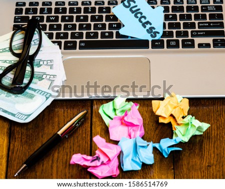 Different objects on wooden office desk. Modern wooden office desk table with laptop keyboard, pen, black glasses, money, crumpled paper and post it with text Success.