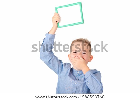 Attractive school boy holds a green frame.Boy smiles and covered mouth shows thumb up.An empty frame with a white background, space for your text.Isolated on white background.
