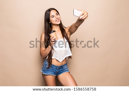 Portrait of pretty young girl taking selfie with mobile phone and waving isolated over beige background