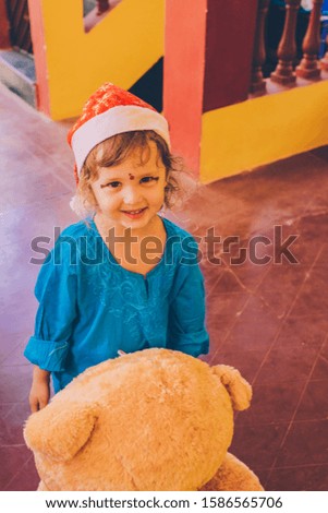 Christmas or New year celebration.  Christmas Travel. Little girl smiling positive in blue dress and santa hat with bear toy. Trip to tropical places on vacation.India.