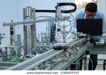 Focus on Gripper which picks up the product from an automated car and programming engineer at the background. Industry 4.0 workshop in an automation education center. Engineer is programming PLC. Royalty-Free Stock Photo #1586609545