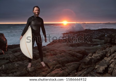 Surfer with surfboard standing on rocks wearing wetsuit with ocean in background and dramatic sunset sky
