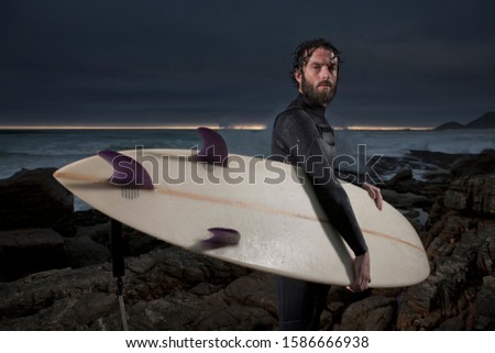 Surfer with surfboard standing on rocks wearing wetsuit with ocean in background and dramatic mood sky