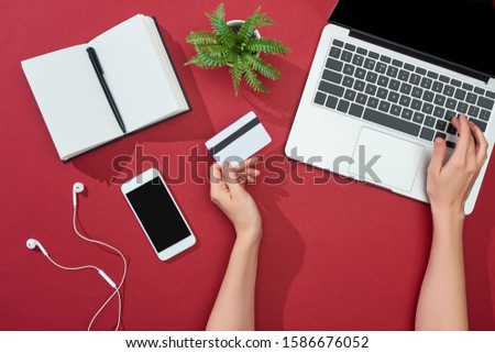 cropped view of woman holding credit card and using laptop near smartphone, earphones, pen, notebook and plant on red background