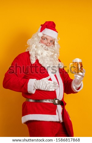 Male actor in a costume of Santa Claus holds in his hands a glass with a light beer and poses on a yellow background