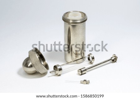 A glass syringe sits on a white countertop next to a cylindrical Nickel-plated metal sterilizer and a needle.   Selective focus. On white background.
