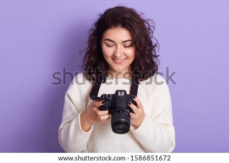 Indoor studio shot of pretty photographer holding her photocamera in both hands, looking at photos, being delighted with results, having pleasant facial expression. Free time activities concept.