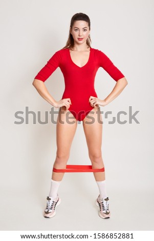 Full length picture of athletic slender model putting her hands on waist, looking directly at camera, having elastic band for fitness around legs, wearing red combidress, white socks and sneakers.