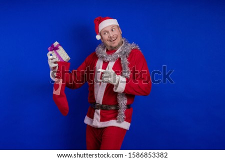Emotional male actor in a costume of Santa Claus holds in his hands a Christmas sock and a box with gifts and poses on a blue background