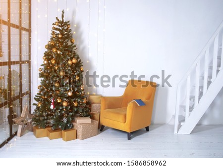 New year's interior. Decorated Christmas tree in the interior of the living room. The blue book is on the yellow chair. White walls in the interior.