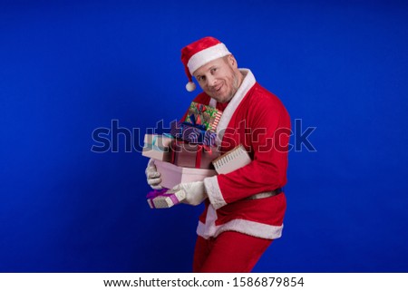 Emotional male actor in a costume of Santa Claus holds gift boxes in his hands and poses on a blue background