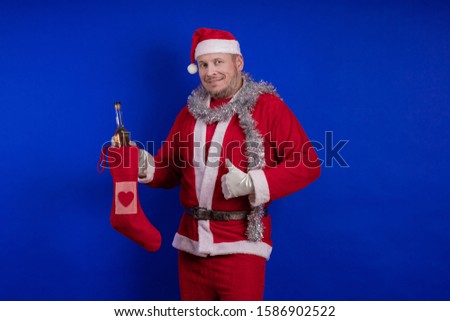 Emotional male actor in a costume of Santa Claus holds in his hands a Christmas sock and a bottle as a gift and poses on a blue background
