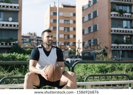 Strong build athlete holds in his hands a basketball while he is waiting sitting on an urban court on a sunny afternoon