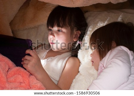 Little girls with an internet tablet under the covers. Baby girls (sisters of two and four years old) are using digital tablet and playing in bed instead of sleeping
