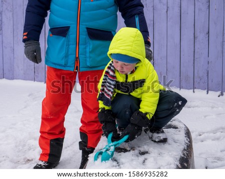 man in bright ski suit with child in yellow jacket is playing with snow in winter yard. baby has snow