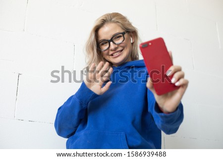 Young woman in glasses takes a selfie on a smartphone and waves her hand. Woman talking via video call.