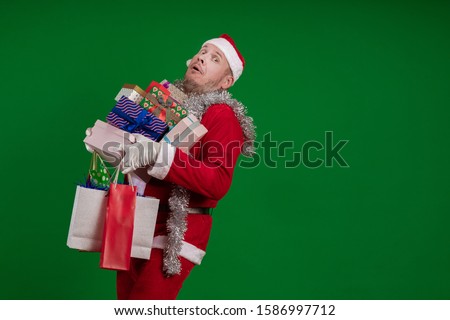 Emotional male actor in a costume of Santa Claus holds a lot of gift boxes and packages in his hands and poses on a green chroma background