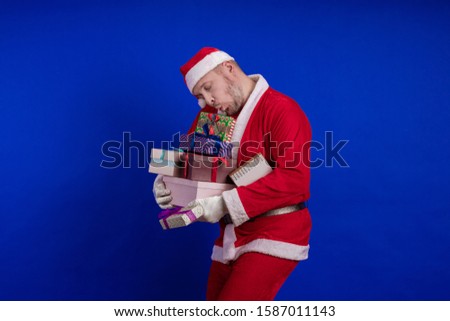 Emotional male actor in a costume of Santa Claus holds gift boxes in his hands and poses on a blue background