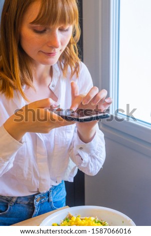 Stock vertical photo of girl taking a picture touching the screen of the mobile to a bowl of food in the kitchen