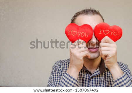 attractive dark-haired man with a smile, with two small red hearts in his hands, covering his eyes with paper heart symbols, standing on a light background. Valentine's day