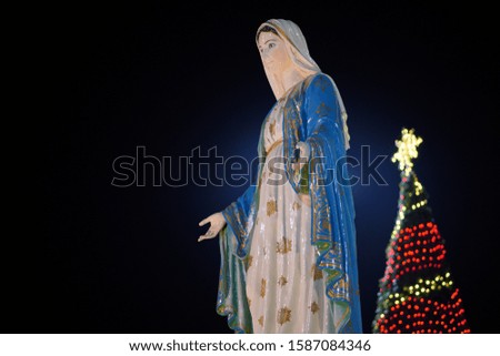 Close up image of mother Mary statue with Christmas tree in background at night, space for text and design