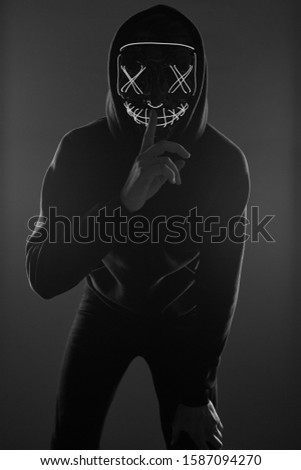 Portrait of an anonymous man in a black hoodie hiding his face behind a scary neon mask. Studio shot.