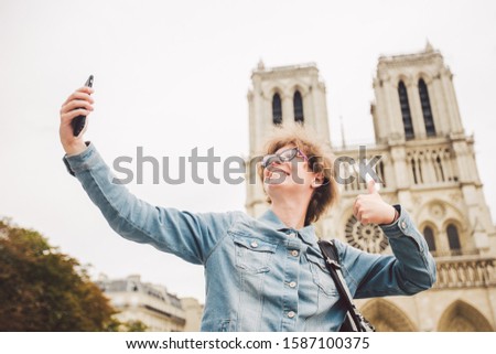 Tourist in Paris making funny selfie near Notre Dame Cathedral. Beautiful young Caucasian tourist woman with backpack in Paris making funny selfie hand holding phone, photo near Notre Dame Cathedral.