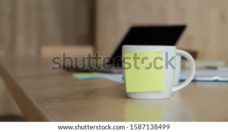A white coffee cup with a blank yellow Sticky Note on the cup. Resting on the desk and laptop is placed on the back.
