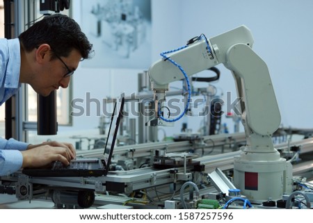 Focus on robotic arm which picks up product from automated car on production line and an engineer working on laptop to programming. Industry 4.0 concept; artificial intelligence in smart factory. Royalty-Free Stock Photo #1587257596