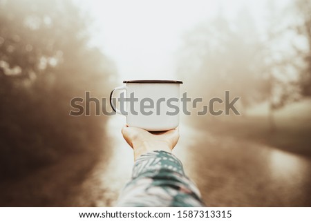 Mockup photo of aluminium mug with coffe in traveler's hand over out of focus river view, tourizm in cold season