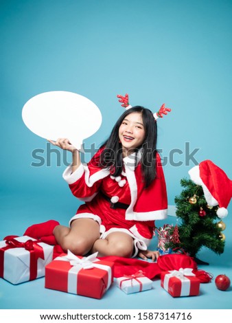 Asian teenager girl wearing Santa Claus costume holding blank speech bubble with gift boxes and Christmas tree on blue background.