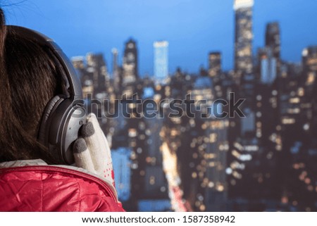 Young woman in a winter jacket and Christmas knitted gloves listening music in the headphones on roof of night New York. Aerial and panorama view of skyscrapers